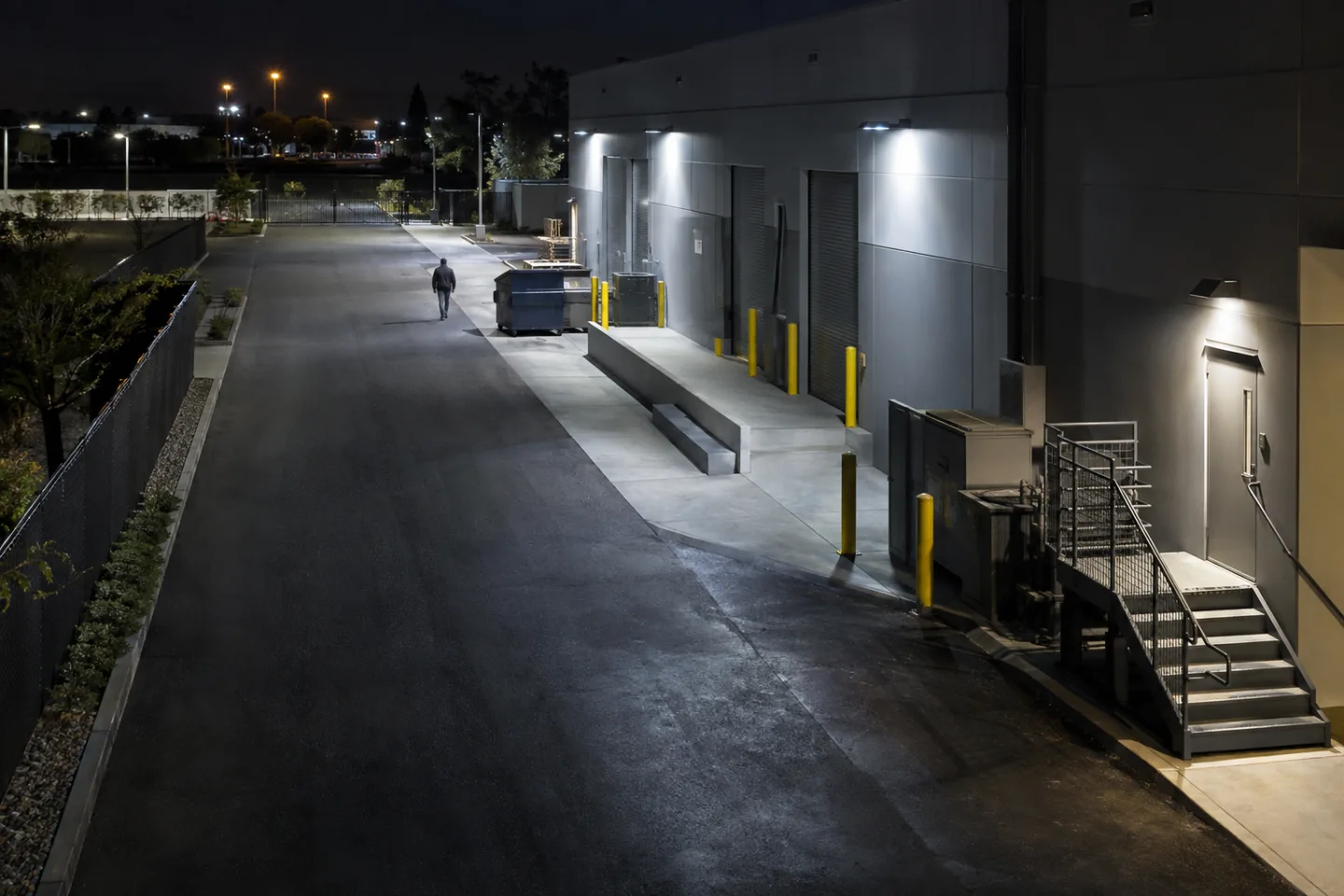 Rear loading entrance of a closed commercial building at night, a distant adult walking near an illuminated access lane, camera mounted high on masonry