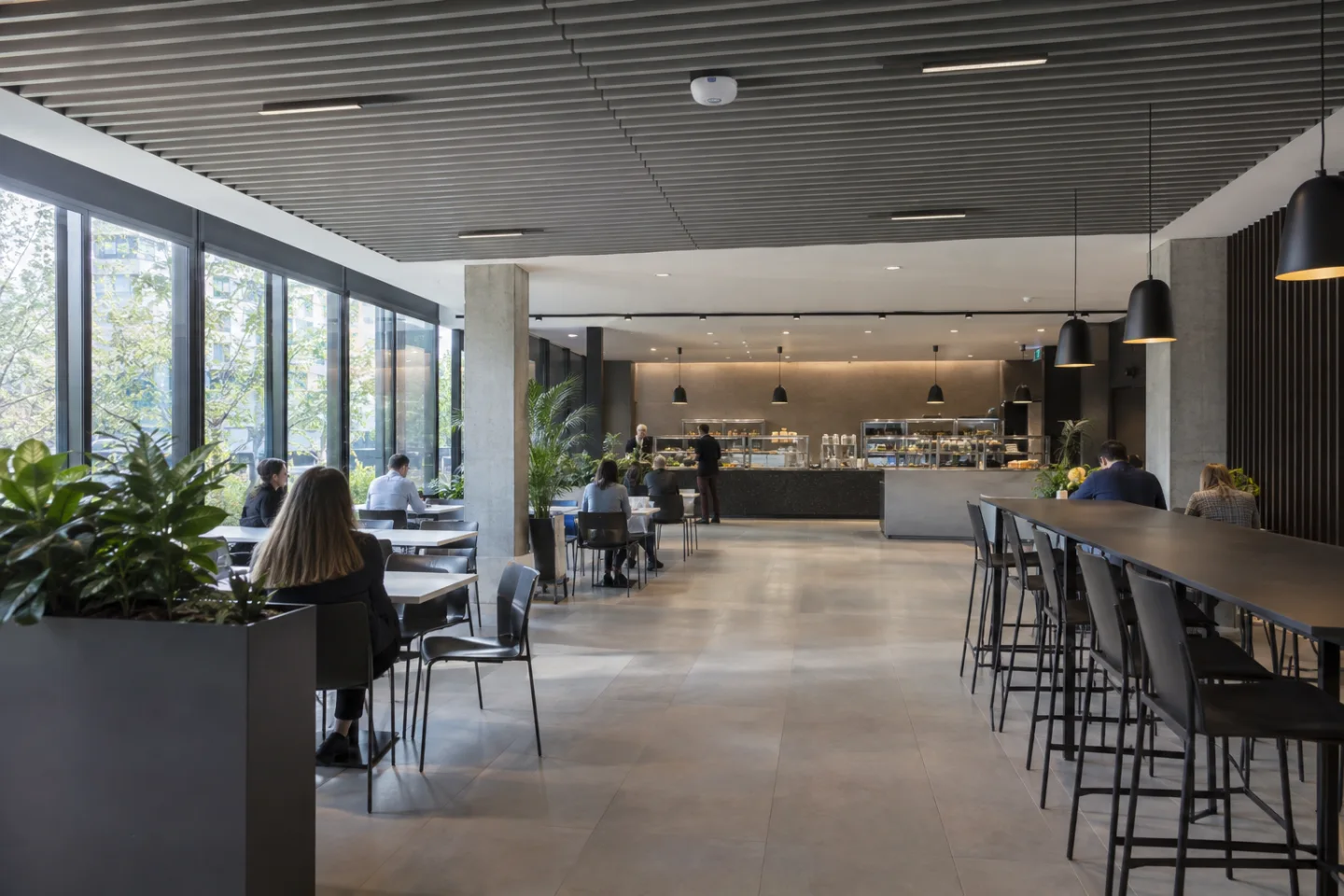 Corporate cafeteria with scattered seated adults and clear walkways, overhead counting sensor, natural window light, wide architectural view